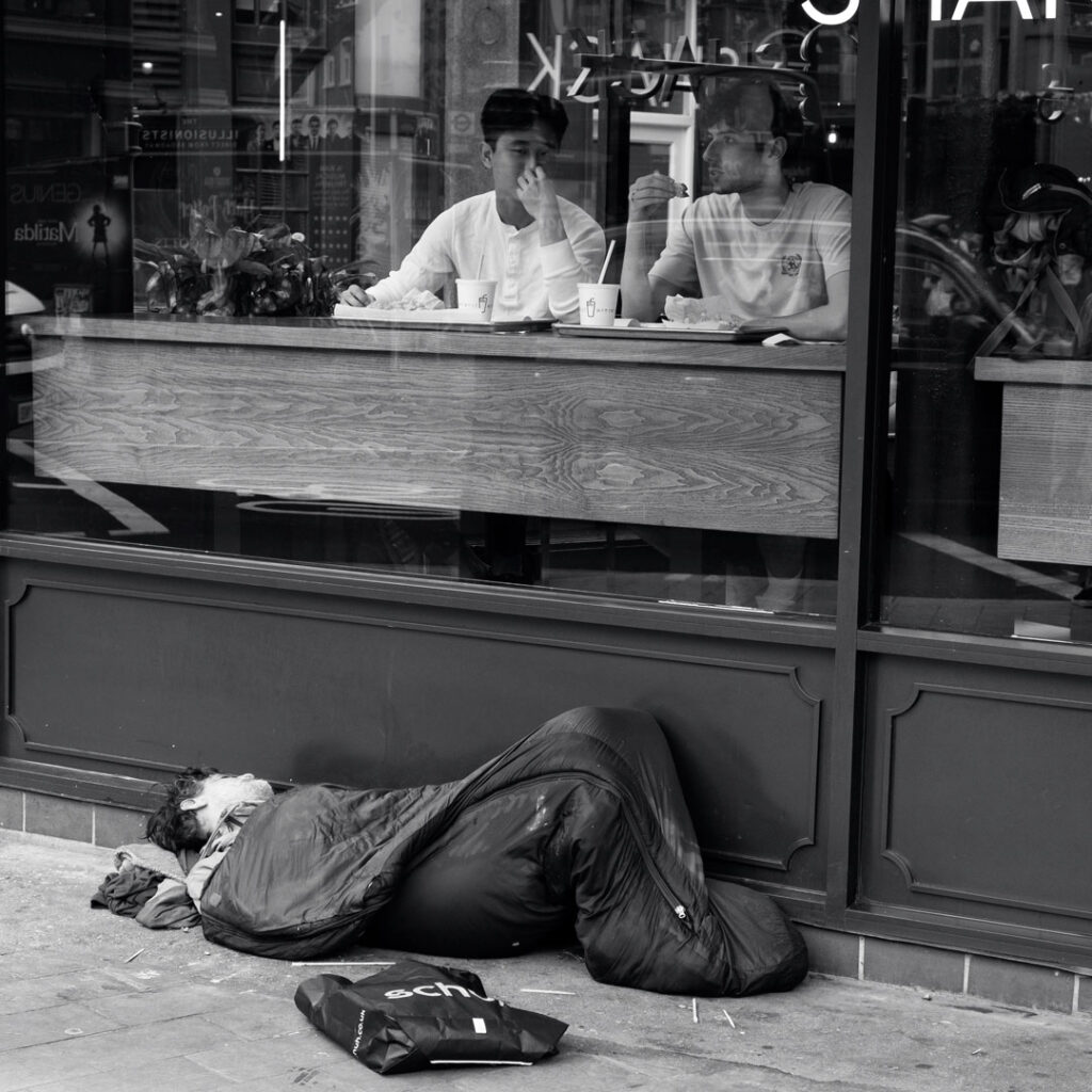 London street photography - Homeless man sleeping outside a restaurant while people eating in window by Adrian Crook